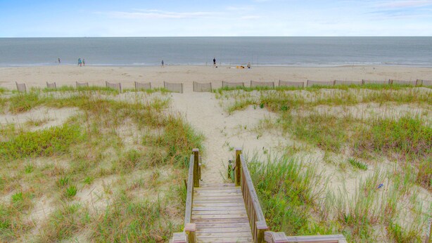 Ein hölzerner Steg führt zu einem Sandstrand mit einer Umzäunung und grasbewachsenen Dünen.