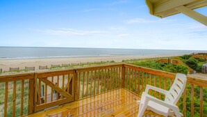 Eine Holzterrasse mit einem weißen Kunststoffstuhl, mit Blick auf einen Strand mit einer Holzpromenade und einem Zaun.