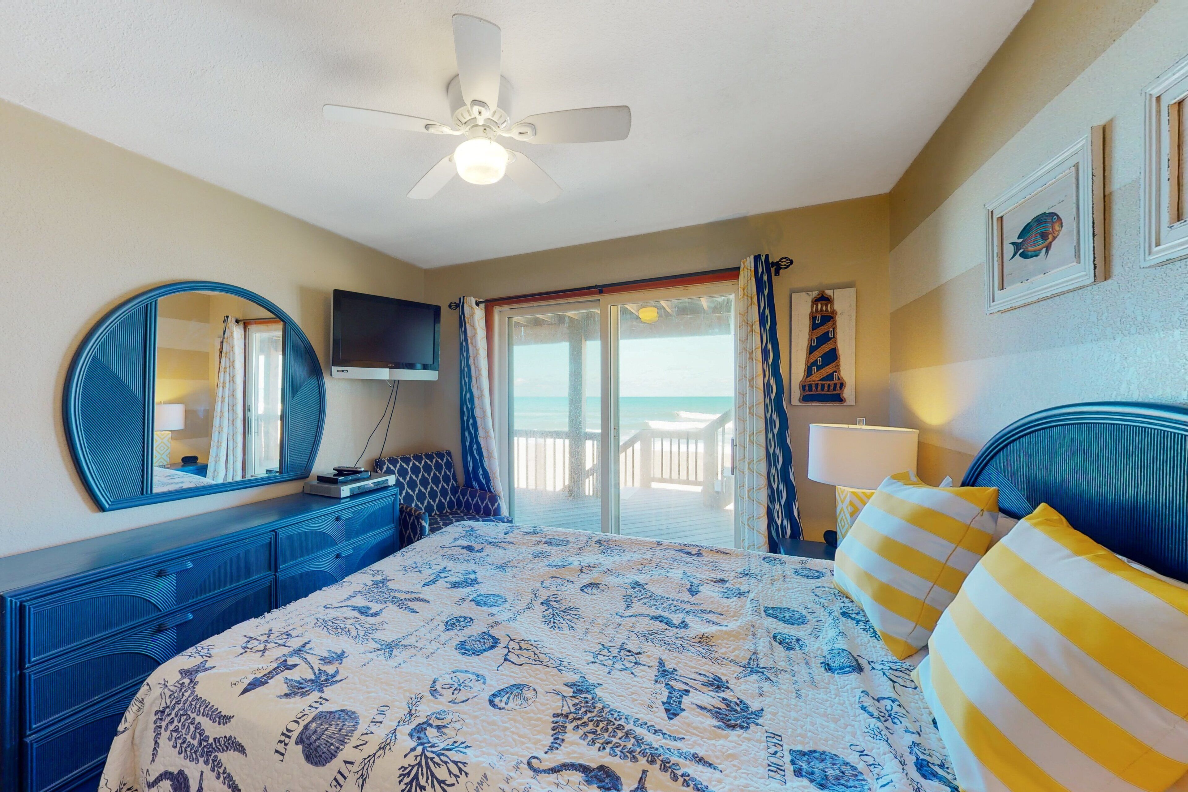 A bedroom with a bed, a ceiling fan, a TV, a large window, and a view of the beach.