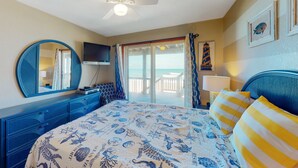 Un dormitorio con cama, ventilador de techo, televisor, ventana grande y vista a la playa.