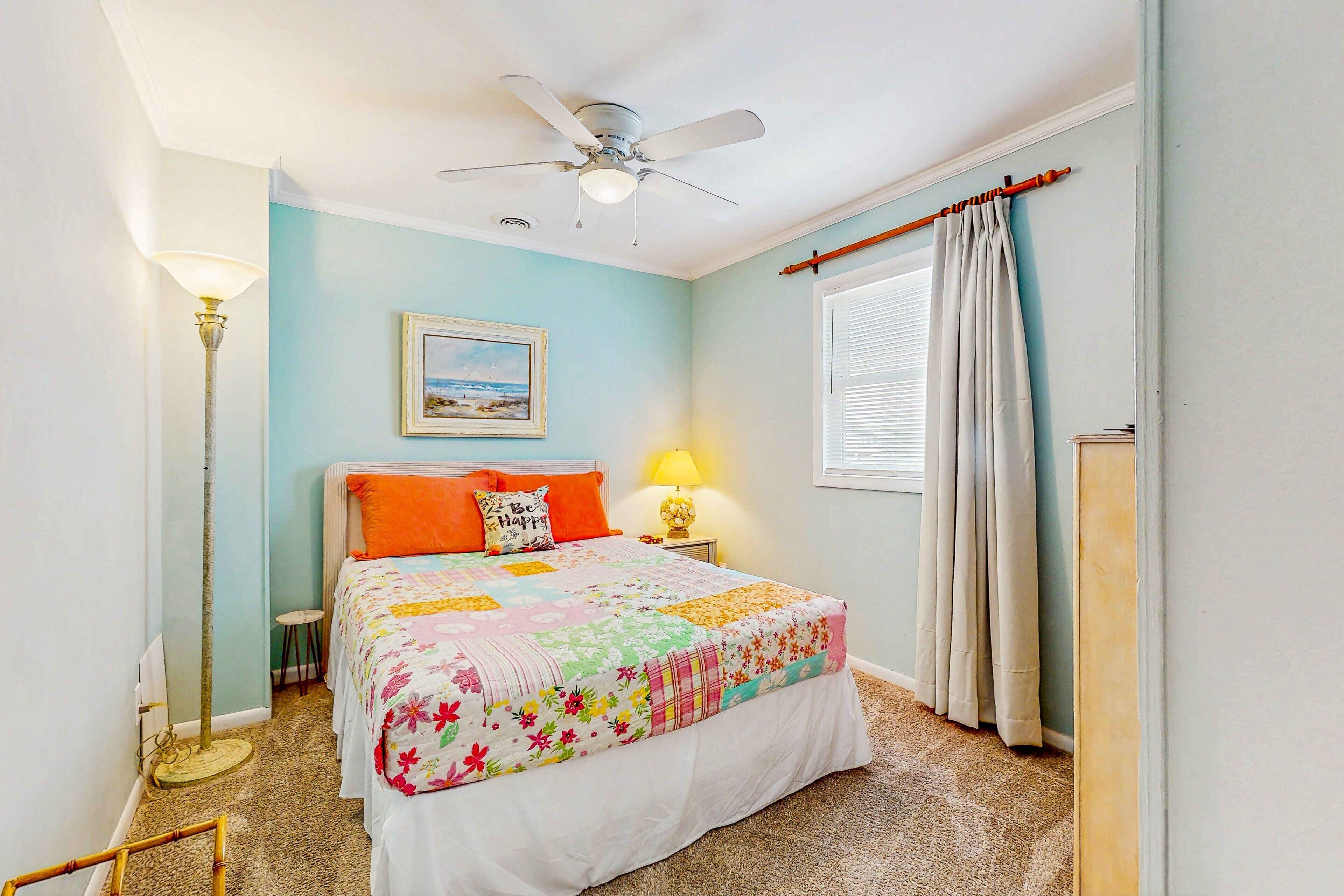 A bedroom with a bed, a ceiling fan, a nightstand with a lamp, a framed picture on the wall, and a window with curtains.