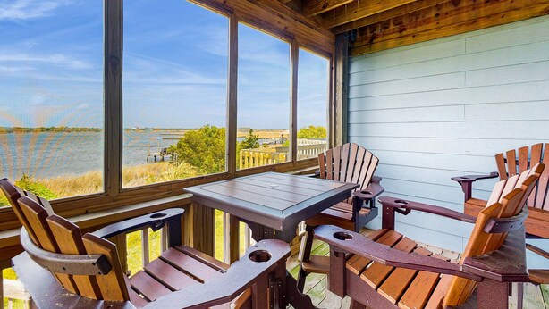 Une terrasse en bois avec une table et des chaises, de grandes fenêtres donnant sur une étendue d’eau.