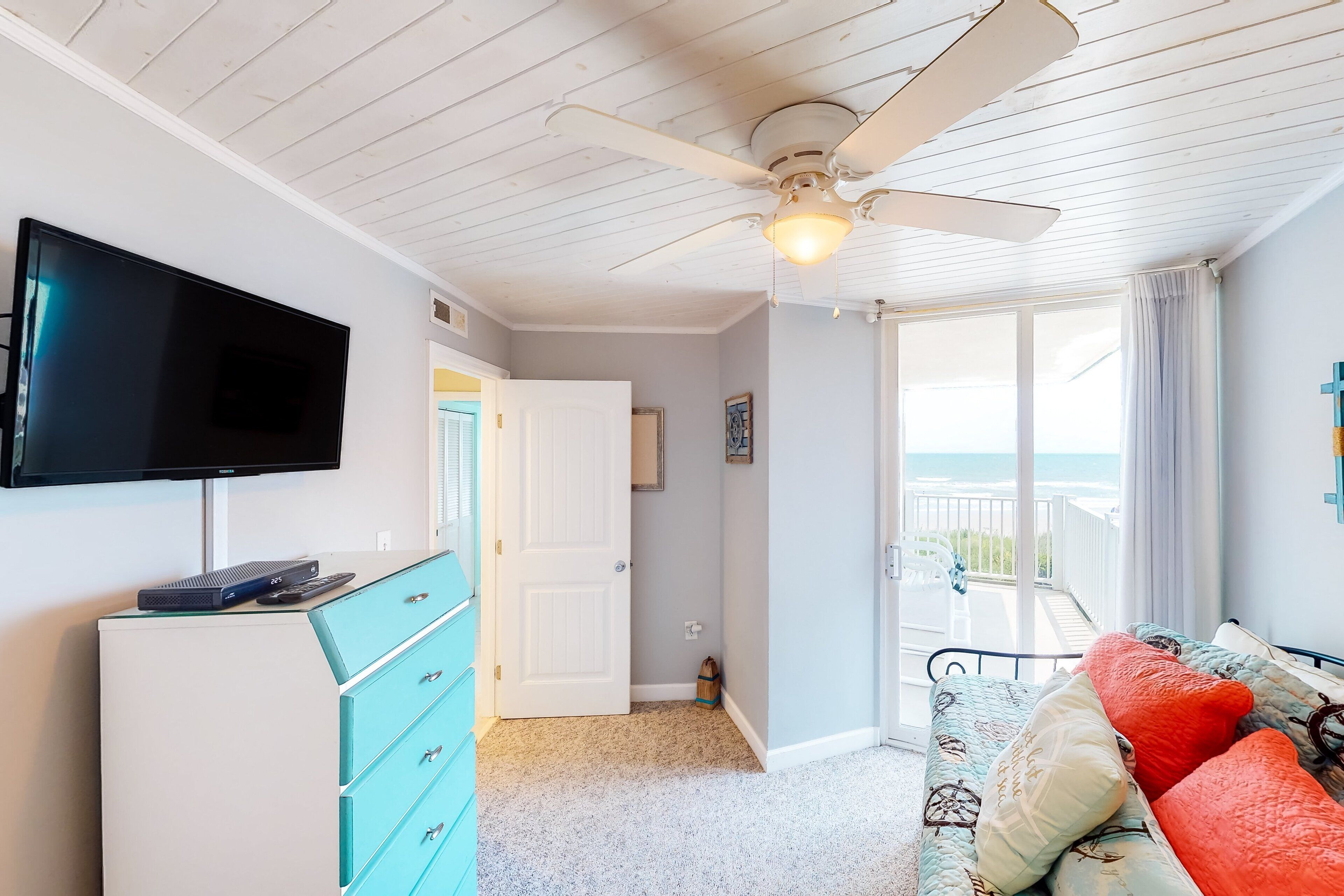 A room with a ceiling fan, a flat-screen TV, a teal dresser, a bed with patterned bedding, and a view of the ocean through a sliding glass door.