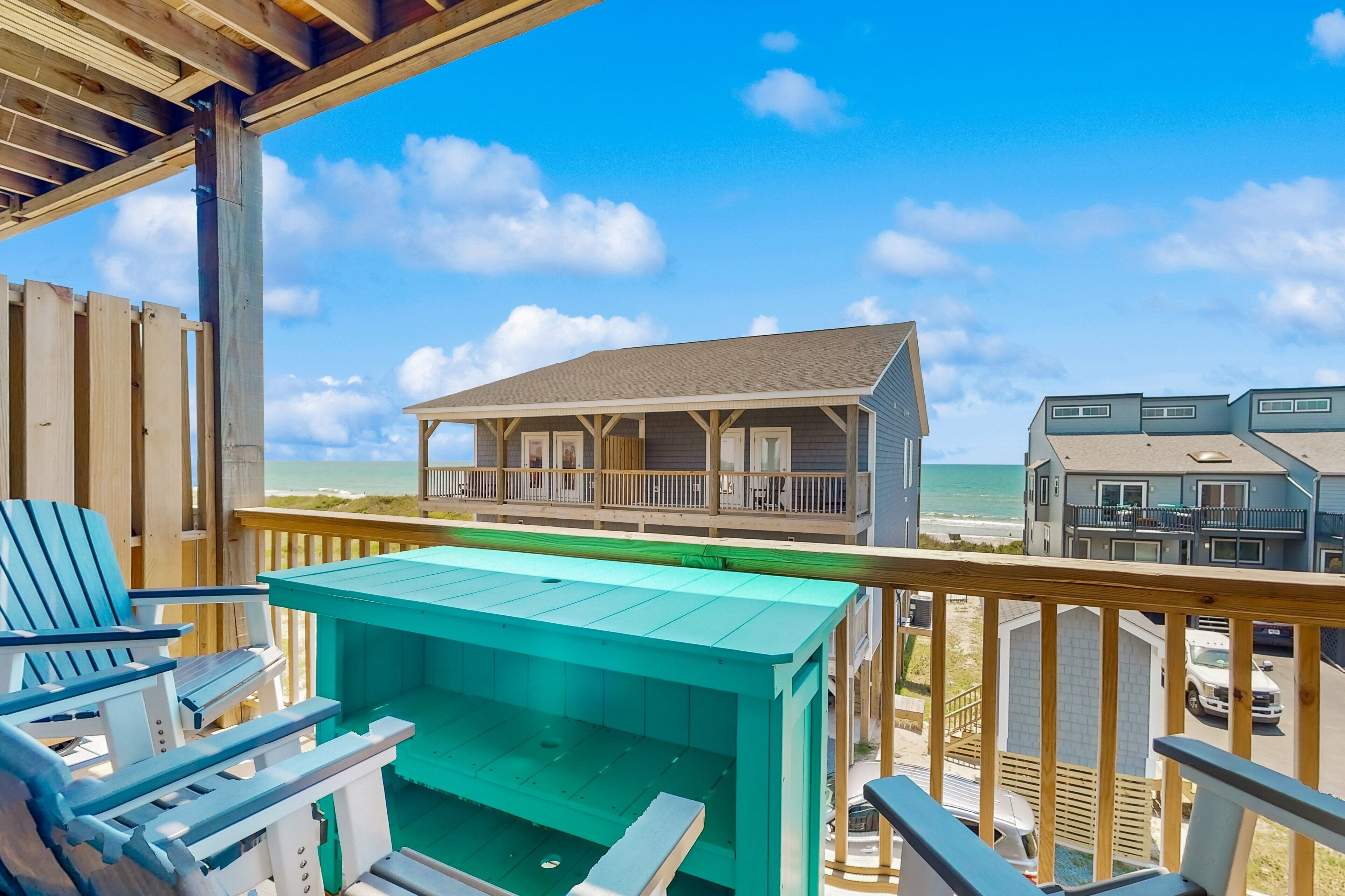 Eine Holzterrasse mit blauen Adirondack-Stühlen und einem grünen Tisch, mit Blick auf ein Strandhaus mit Balkon.