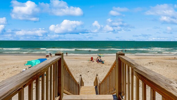 Un escalier en bois menant à une plage, avec un ciel dégagé et une vue sur l’océan.