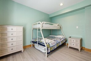 Un lit superposé avec une courtepointe, une commode avec de nombreux tiroirs et une table de chevet dans une pièce aux murs de couleur claire et au plancher en bois.
