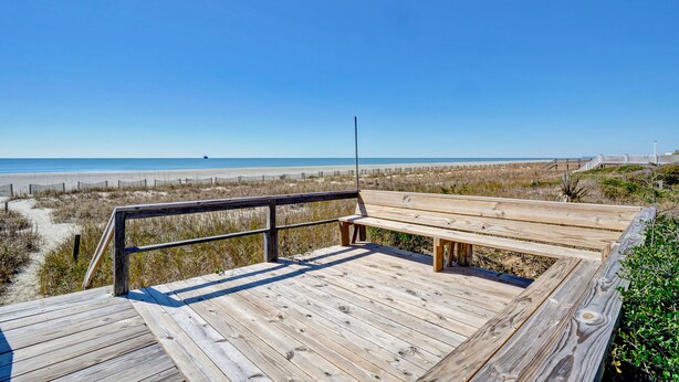 Een houten terras met een bankje met uitzicht op een strand en duinen.