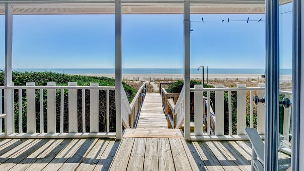 Une terrasse en bois menant à un point de vue sur la plage, avec une balustrade blanche et un ciel dégagé.