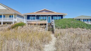 Une propriété située en bord de mer, dotée d’une terrasse en bois, d’un escalier menant à l’entrée et offrant une vue dégagée sur l’océan.