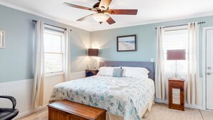 Un dormitorio con cama, ventilador de techo, mesita de noche con lámpara, banco de madera y ventana con cortinas.