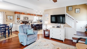 Amplia sala de estar con un sillón gris grande, un comedor con mesa y sillas de madera, una cocina con armarios blancos y una escalera que sube a otro nivel.