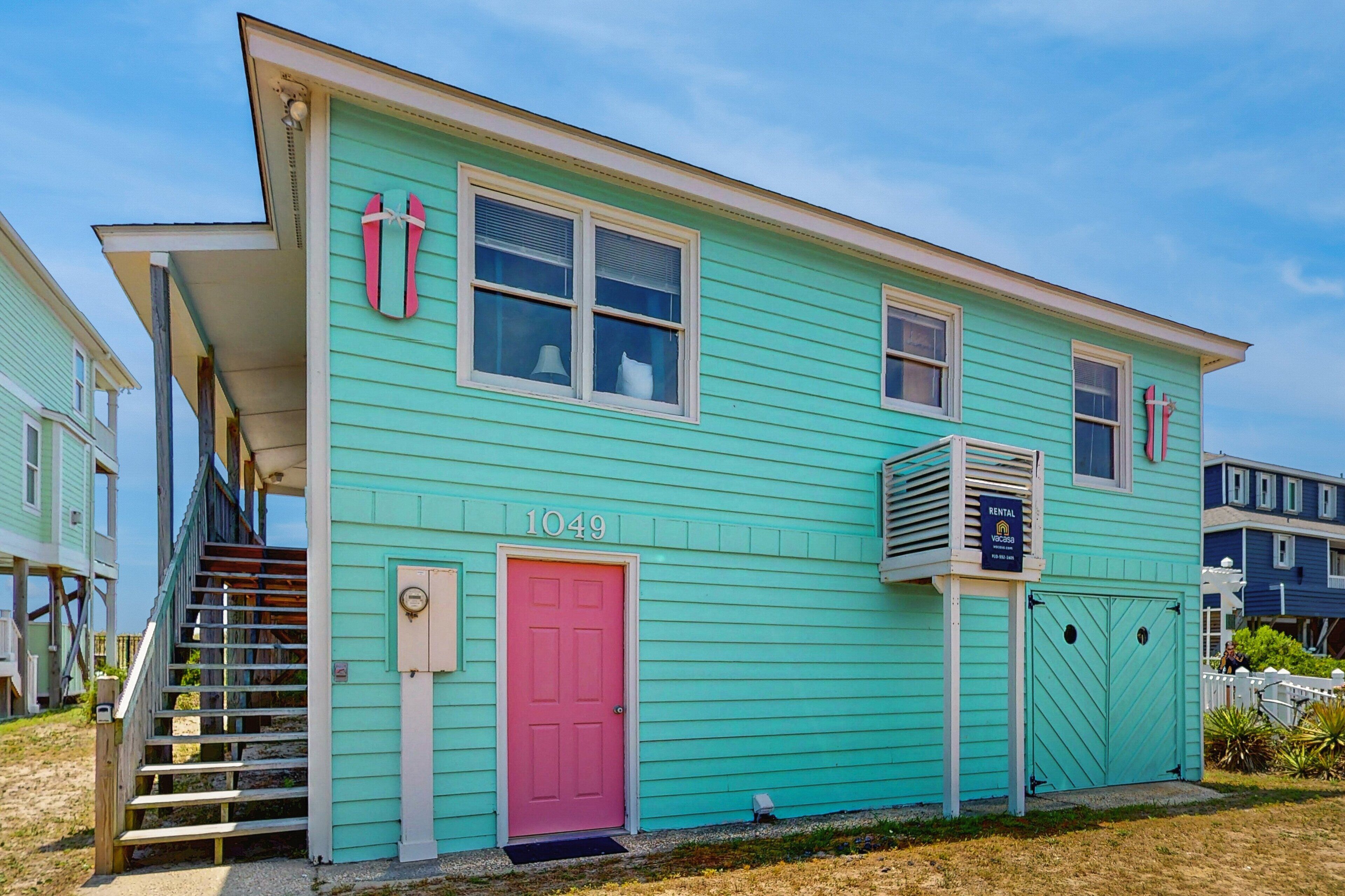 A turquoise two-story house with a pink door, numbered 1049, and a visible staircase leading to the entrance.