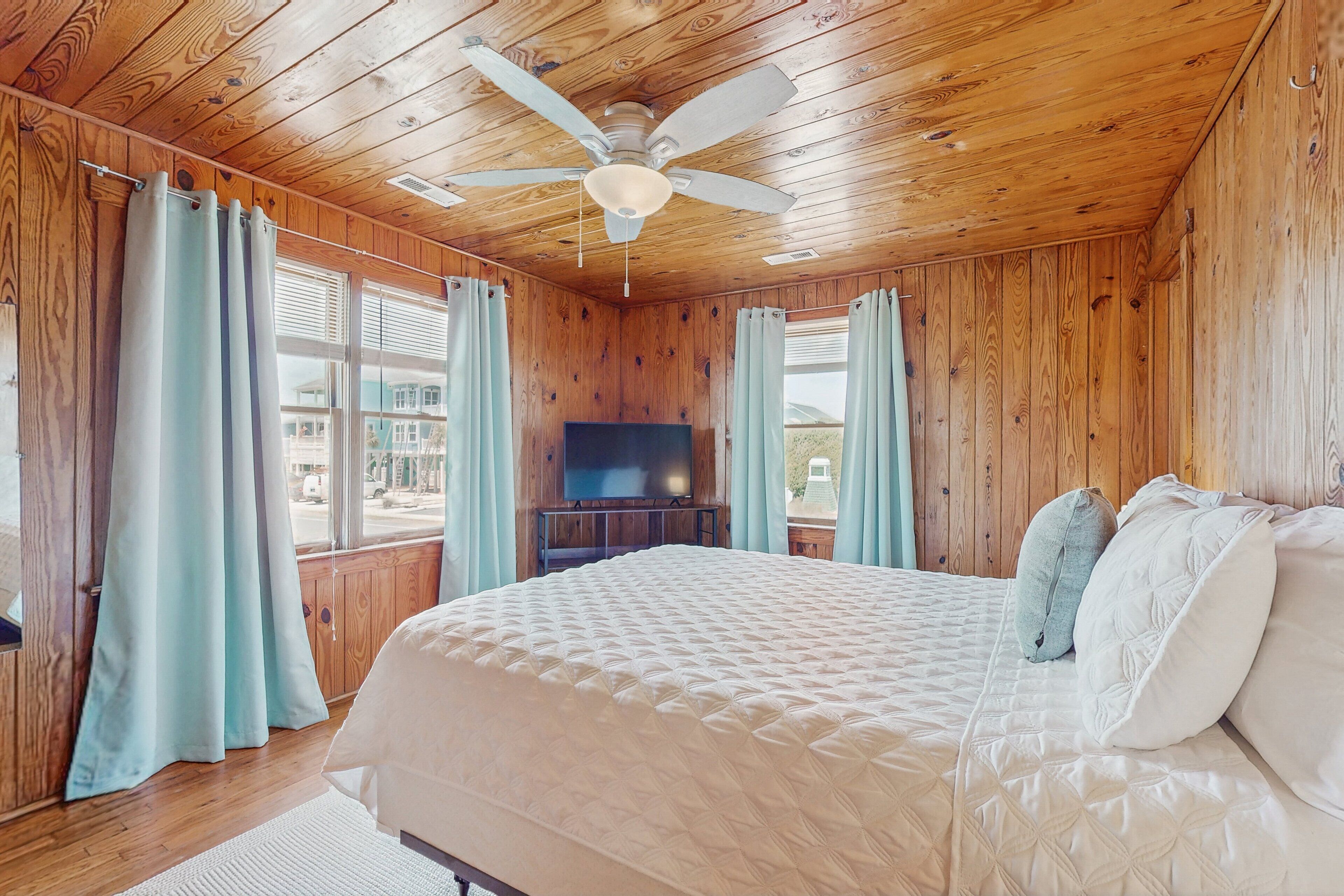 A wooden-paneled bedroom with a bed, curtains, a ceiling fan, and a TV.