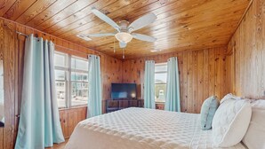 A wooden-paneled bedroom with a bed, curtains, a ceiling fan, and a TV.