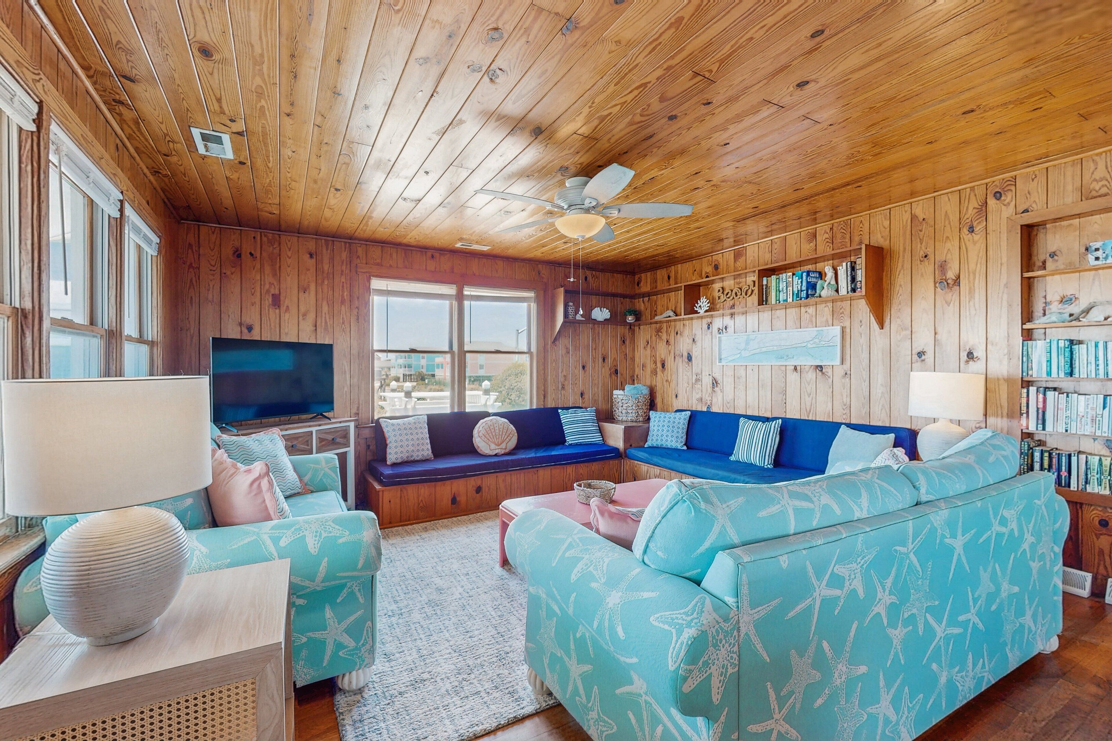A cozy living room with wooden walls and ceiling, a large window, a ceiling fan, and a bookshelf filled with books.