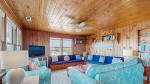 A cozy living room with wooden walls and ceiling, a large window, a ceiling fan, and a bookshelf filled with books.