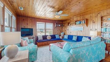A cozy living room with wooden walls and ceiling, a large window, a ceiling fan, and a bookshelf filled with books.