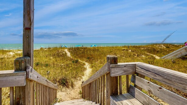 A wooden boardwalk leading to a beach with dunes and a clear sky.