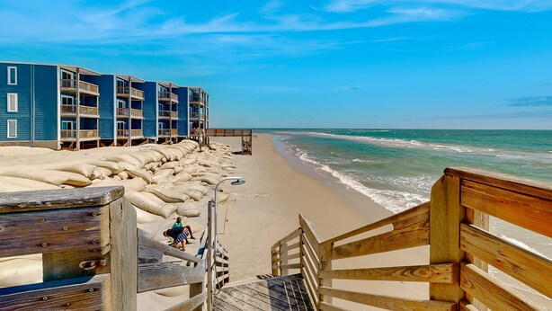 Una escalera de madera que baja a una playa con sacos de arena y un edificio azul al fondo.