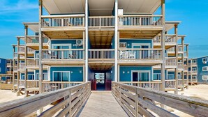 Un edificio frente a la playa con varios pisos, balcones y una terraza de madera que lleva a la entrada.
