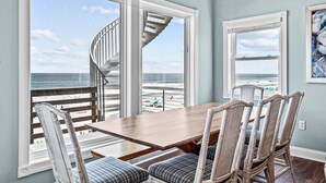 Un espace repas avec une table en bois et des chaises en osier, de grandes fenêtres offrant une vue sur la plage et l’océan, et un escalier en colimaçon menant à un niveau supérieur.