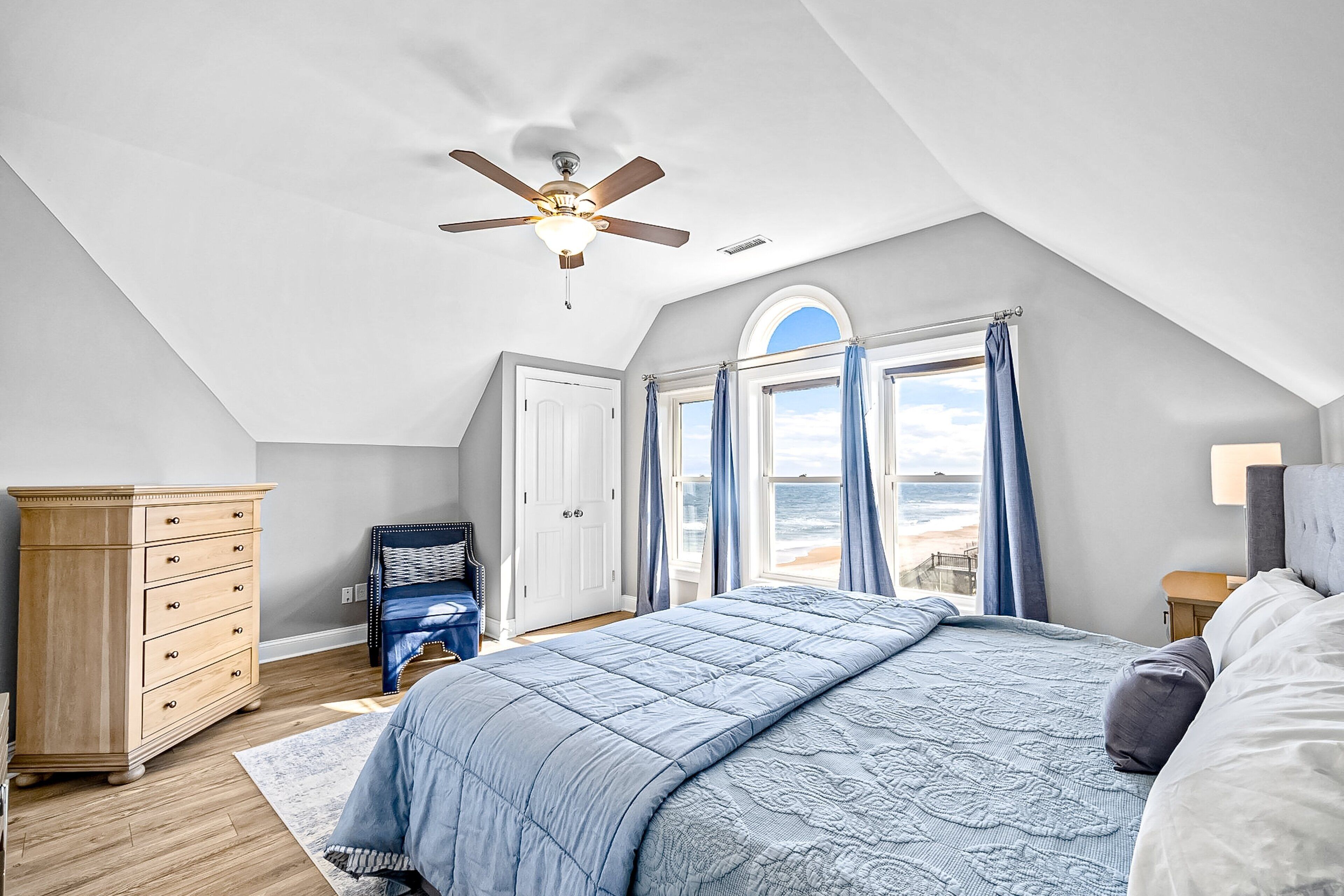 Une chambre avec un grand lit, un ventilateur de plafond, une commode et une vue sur l’océan par les fenêtres.