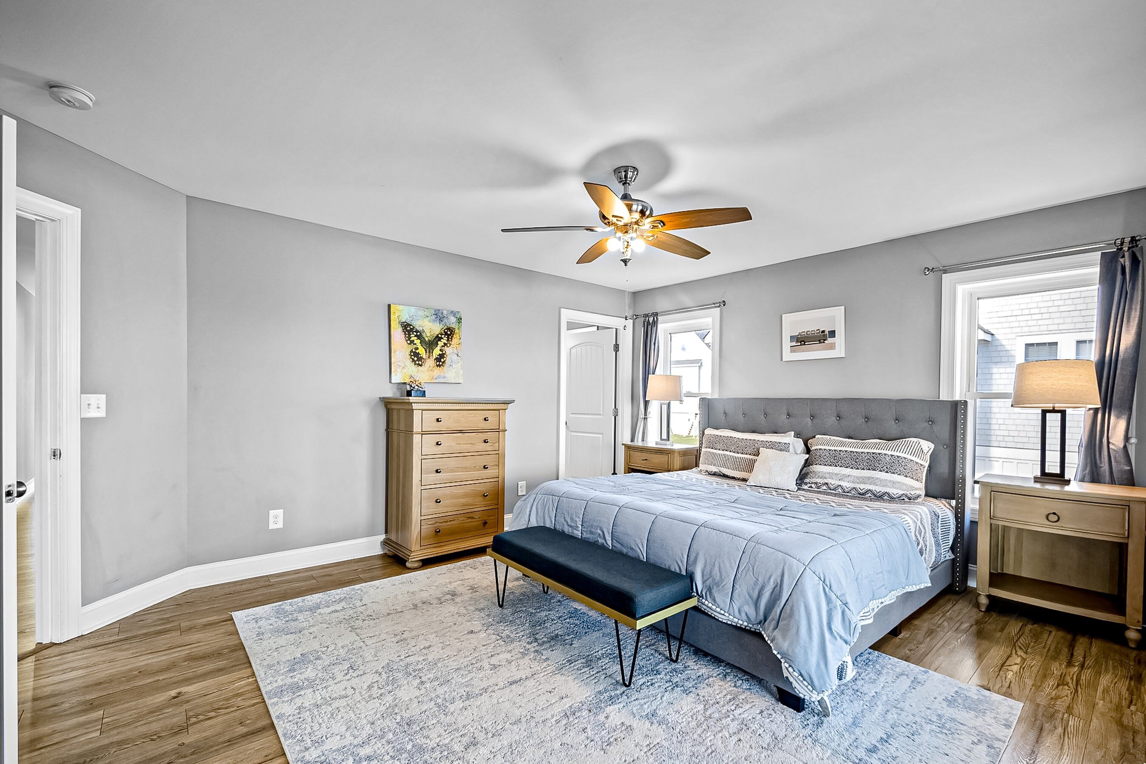 A bedroom with a bed, a bench, a dresser, a ceiling fan, and a window with curtains.