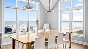 Un comedor con una mesa de madera y sillas blancas, ventanas amplias con vista al océano, e iluminación colgante.