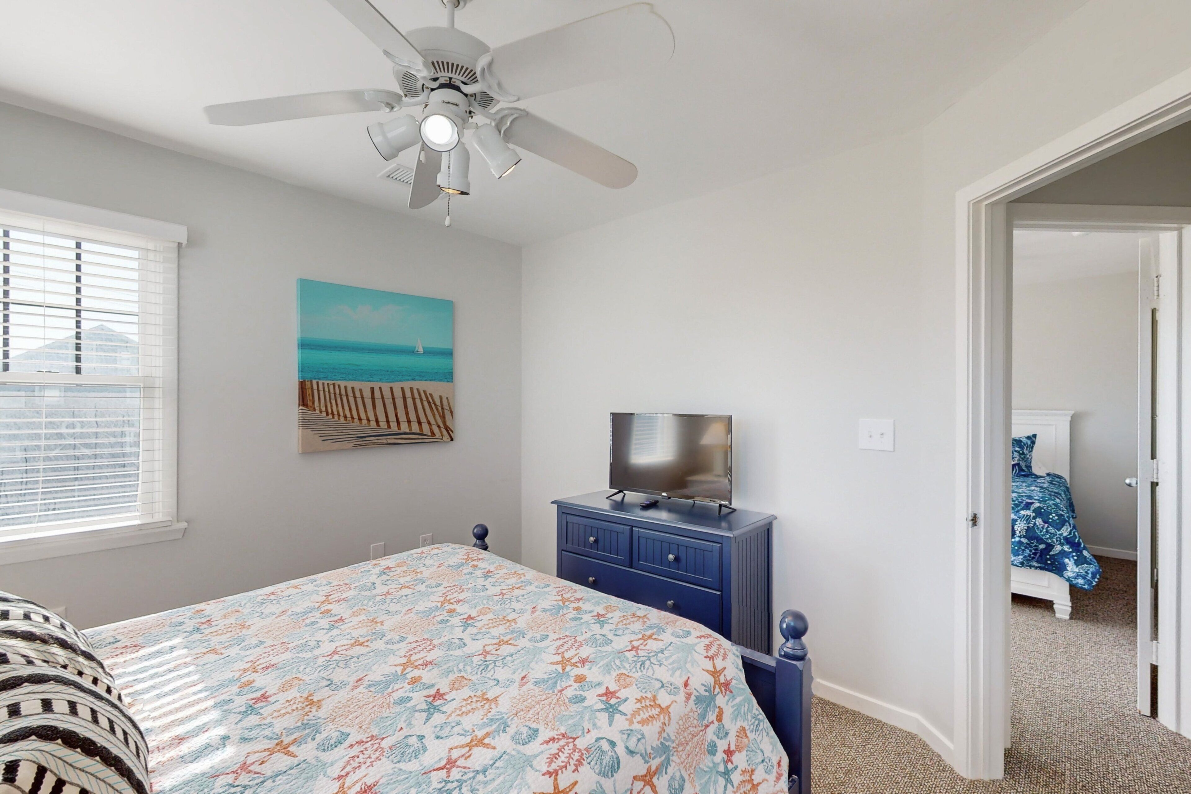 A bedroom with a bed, a ceiling fan, a TV on a dresser, a painting of a beach, and a door leading to another room.