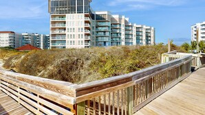 A modern multi-story building with balconies, a wooden boardwalk, and a beach in the background. - Oceanfront condo with pool, sauna, gym, game room, on-site restaurants, balcony (North Topsail Beach)