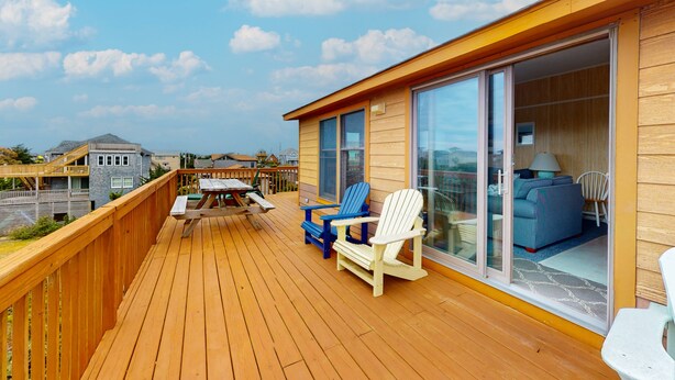 A wooden deck with a table and chairs, a sliding glass door leading to a room with a sofa and lamp, and a view of other buildings. - Spacious dog-friendly house with shared pool, private hot tub, & central AC (Avon)