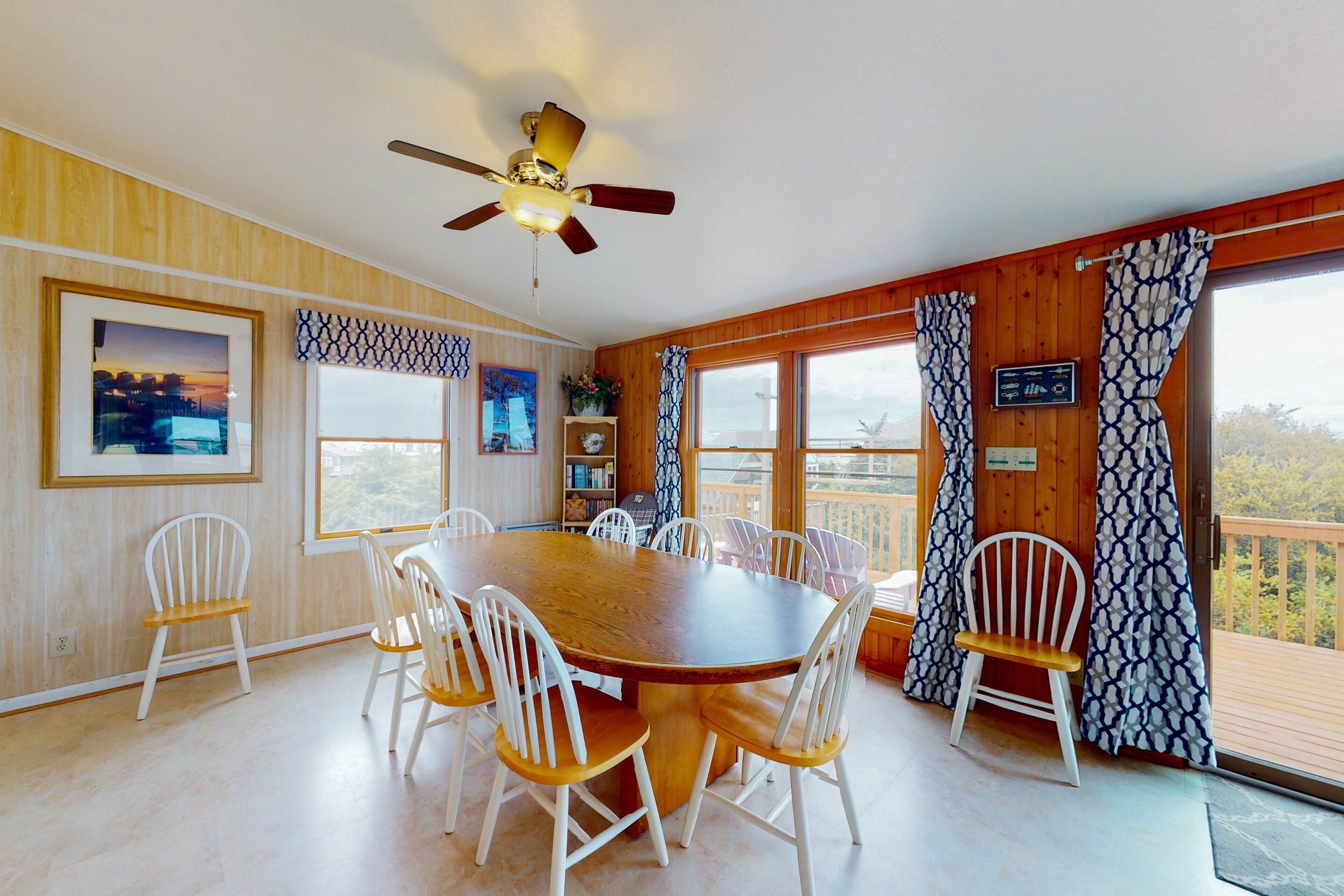 A dining area with a wooden table and white chairs, a ceiling fan, and large windows with patterned curtains.