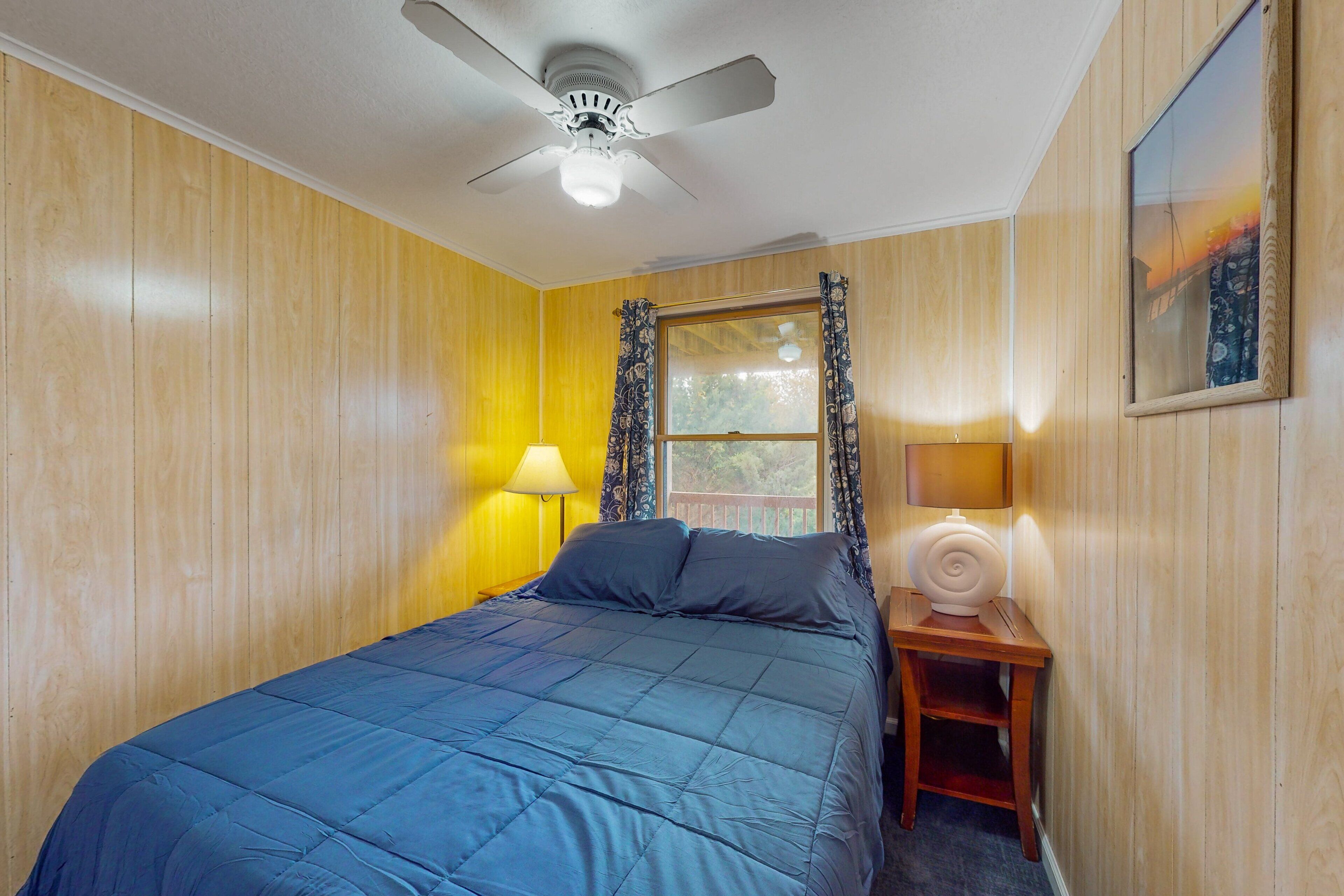 A bedroom with a bed, a ceiling fan, a window with curtains, and a nightstand with a lamp.