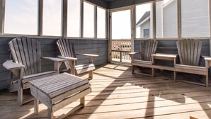A sunlit wooden deck with Adirondack chairs and tables, large windows, and a view of a house. - Beautiful dog-friendly home overlooking the beach with hot tub, WiFi, & AC (Salvo)