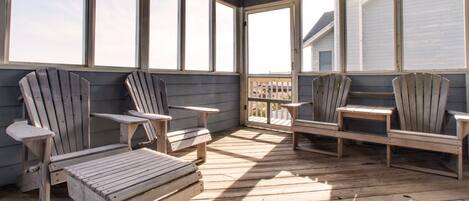 A sunlit wooden deck with Adirondack chairs and tables, large windows, and a view of a house.