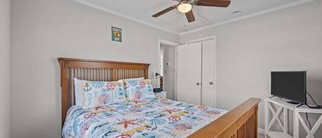 Un dormitorio con cama, ventilador de techo, televisor sobre un mueble y una puerta.