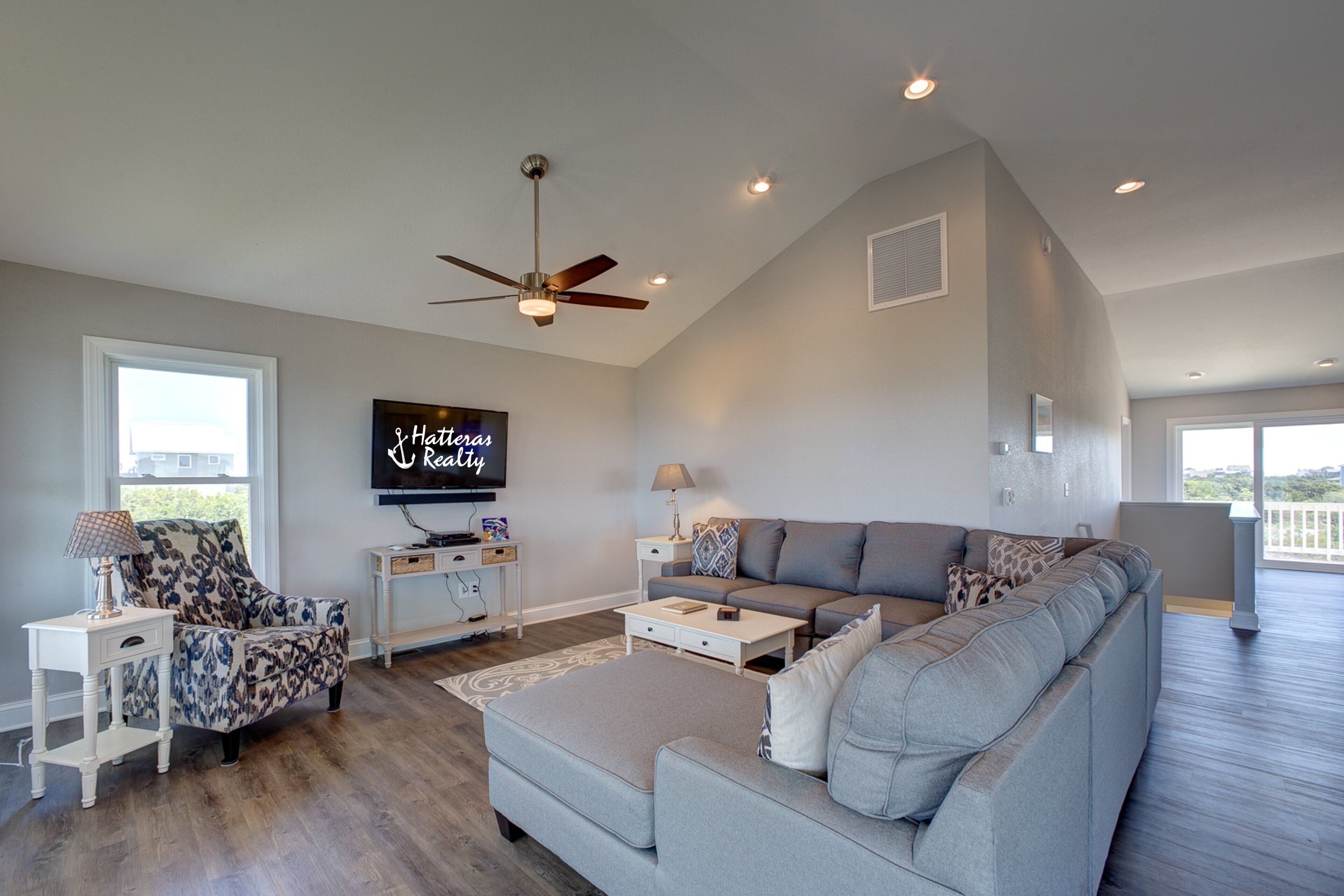 A spacious living room with a grey sofa, patterned armchair, and a flat-screen TV mounted on the wall.