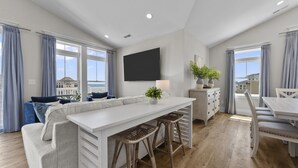 A modern living room with a white sofa, a dining table with bar stools, and a flat-screen TV mounted on the wall. - Oceanview Home with private pool and close beach access (North Topsail Beach)