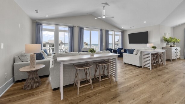 A modern living room with a white sofa, a dining table with bar stools, a ceiling fan, and large windows with curtains. - Oceanview Home with private pool and close beach access (North Topsail Beach)