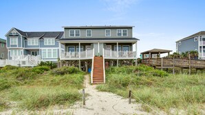 En ejendom ved stranden med en central trappe, der fører op til en balkon, omgivet af sand og vegetation.