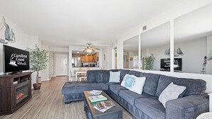 A modern living room with a grey sectional sofa, a coffee table with magazines, a television on a wooden stand, and a large mirror reflecting the room. - Oceanfront condo with shared pool, shared sauna and beach access (North Topsail Beach)