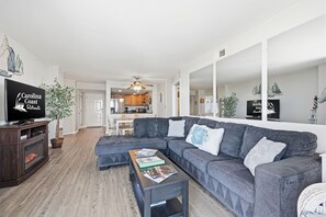 A modern living room with a grey sectional sofa, a coffee table with magazines, a television on a wooden stand, and a large mirror reflecting the room.