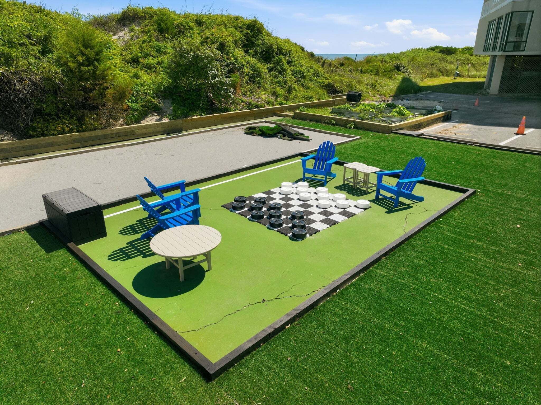 A chessboard set up on artificial grass with blue chairs and a table, surrounded by greenery and a road.