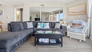 A living room with a grey sectional sofa, a white wicker chair, a coffee table with magazines, and a painting on the wall. - Oceanfront condo with shared pool, shared sauna and beach access (North Topsail Beach)