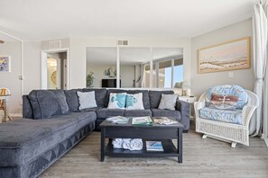 A living room with a grey sectional sofa, a white wicker chair, a coffee table with magazines, and a painting on the wall.