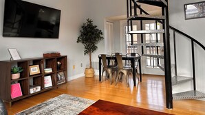 Ein modernes Wohnzimmer mit Flachbildfernseher, Holzböden, einem Essbereich mit Stühlen, einer Treppe und einem großen Fenster.
