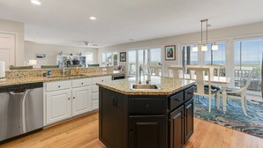 Eine moderne Küche mit Granitwerkplatten, weißen Schränken und einer großen Kochinsel mit Spüle. Ein Essbereich mit Tisch und Stühlen bietet einen Blick auf das Meer durch große Fenster.