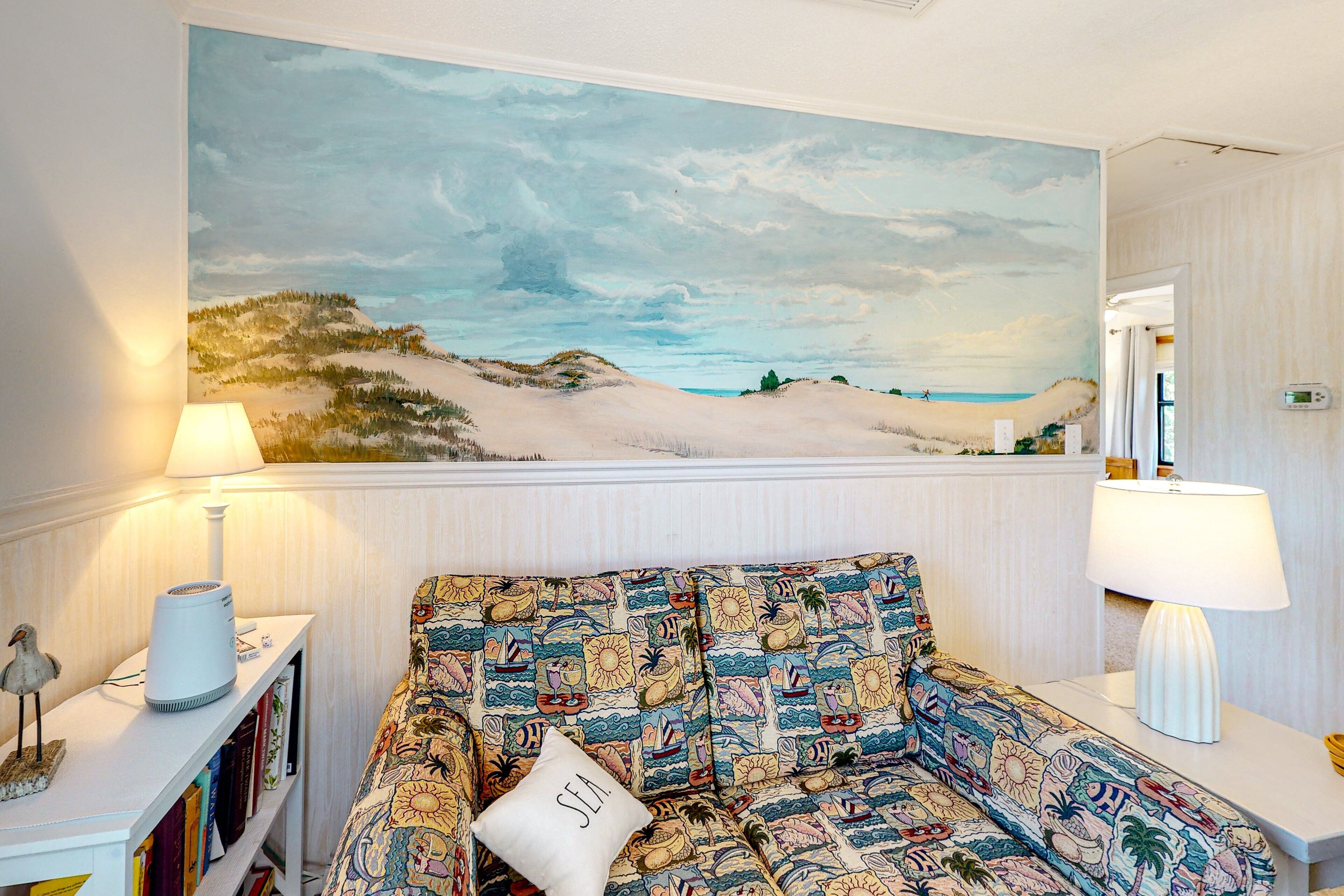 A room with a large beach landscape painting, a sofa with a patterned cover, and a side table with a lamp.