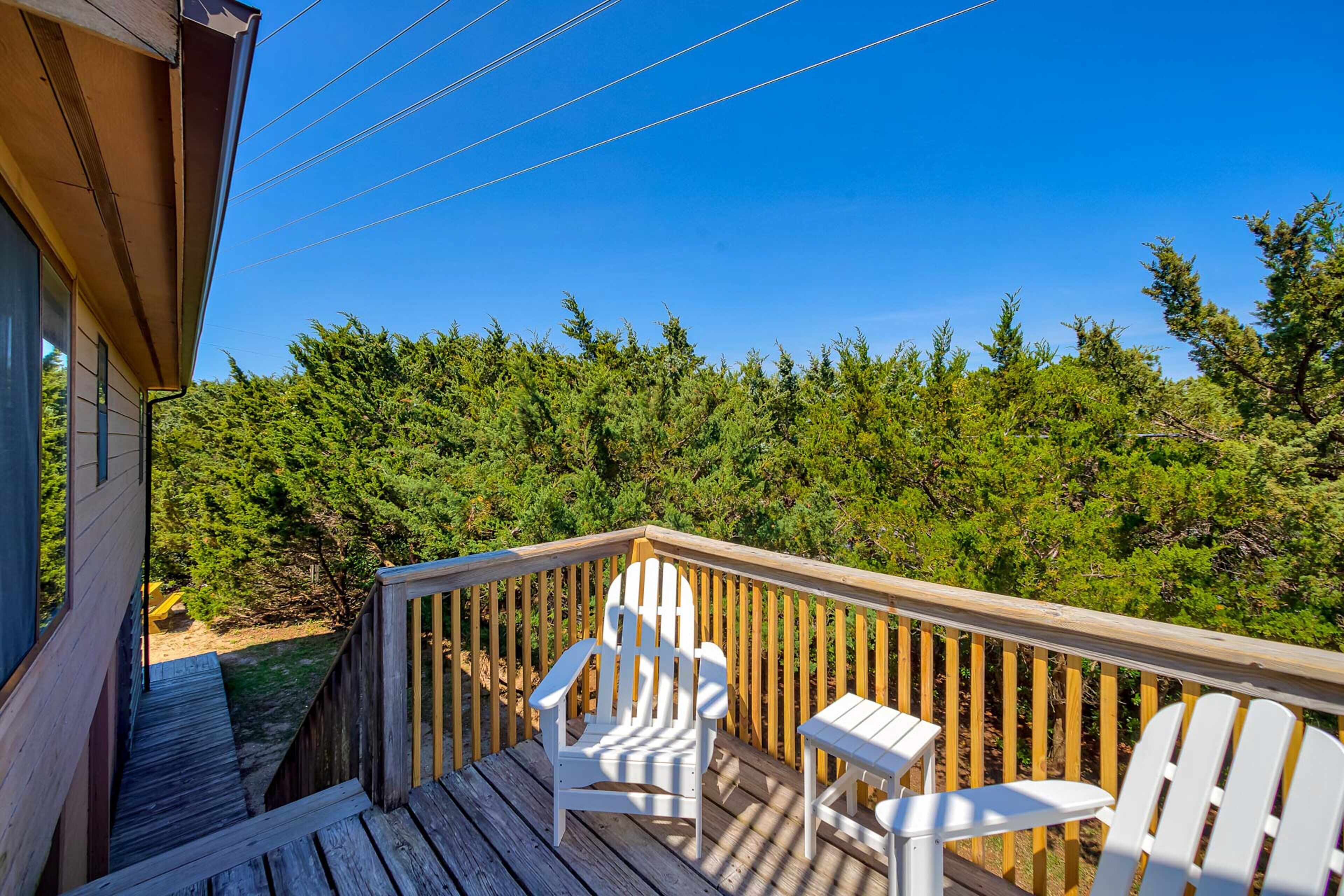 A wooden deck with white Adirondack chairs and a small table, overlooking a dense forest.