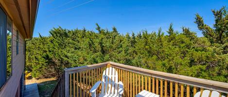 A wooden deck with white Adirondack chairs and a small table, overlooking a dense forest.